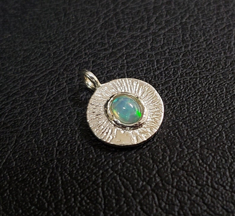 May include: A silver pendant with a round, textured frame and a light blue opal gemstone in the center.