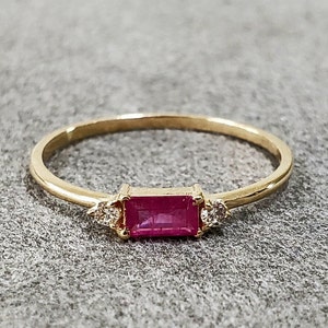 Op de afbeelding: Een gouden ring met een roze rechthoekige edelsteen en twee kleinere heldere edelstenen aan weerszijden.
