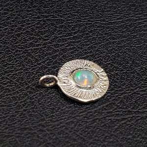 May include: A silver pendant with a textured circular design and a small, iridescent opal gemstone in the center.