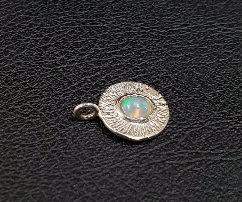 May include: A silver pendant with a textured circular design and a small, iridescent opal gemstone in the center.