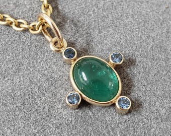 May include: Gold-toned necklace with a pendant featuring an oval, emerald-green gemstone. The pendant is accented with four small, round, blue gemstones. The necklace chain is also gold-toned.