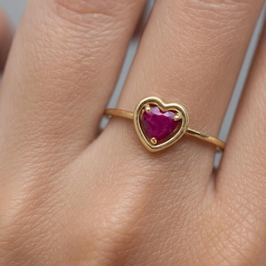Puede incluir: Un anillo de oro con una piedra preciosa roja oscura en forma de corazón. La piedra preciosa está engastada en un marco de oro en forma de corazón. El anillo está en un dedo, mostrando el diseño y el color de la joya.