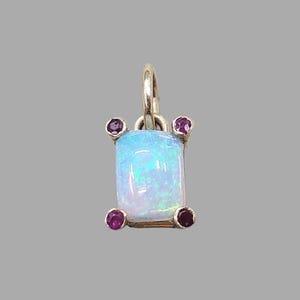 May include: A silver pendant featuring a rectangular opal gemstone with iridescent blue and green hues. Four small, round, deep red gemstones are set around the opal. The pendant has a small loop at the top for a chain.
