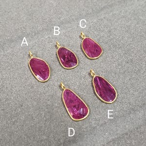 May include: Five gold-plated pendants with irregular, faceted, deep pink gemstones. Each pendant has a small loop at the top for a chain. The gemstones are set in a gold bezel. The pendants are labeled A, B, C, D, and E.