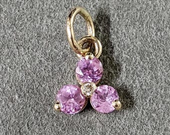 May include: A gold charm with three pink gemstones and a small diamond in the center. The charm has a loop at the top for attaching to a necklace or bracelet.