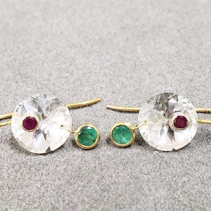 Puede incluir: Un par de pendientes de oro con piedras redondas transparentes y una piedra roja más pequeña en el centro. Cada pendiente tiene una piedra verde colgante.