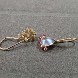 May include: A pair of gold earrings with a teardrop-shaped, iridescent moonstone and small red gemstones. The earrings have a floral design and a simple hook closure. The jewelry is displayed on a gray surface.