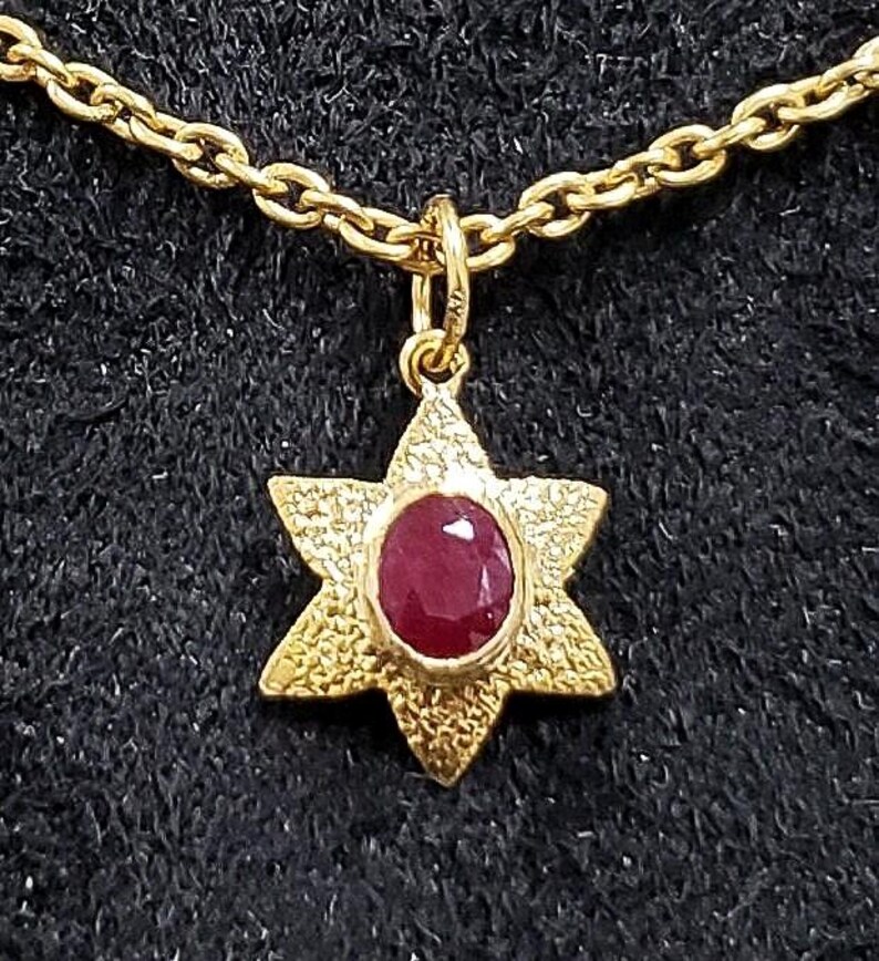 May include: A gold-colored necklace featuring a pendant in the shape of a six-pointed star. The star has a textured surface and a central oval-shaped, deep red gemstone. The chain is also gold-colored and has a link design.