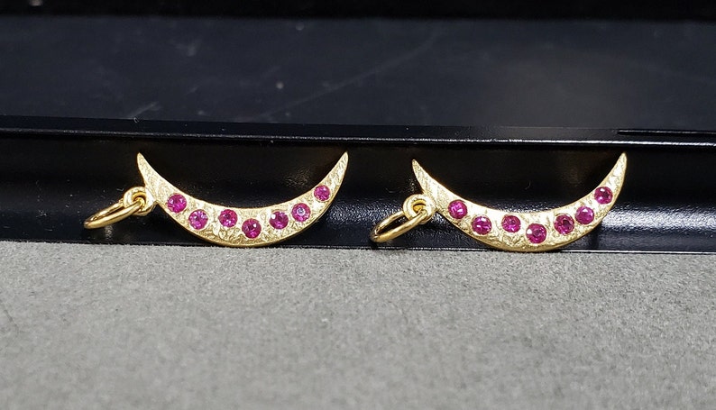 May include: Two gold crescent moon charms with pink gemstones set along the edge.