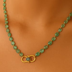 Puede incluir: Un collar dorado con piedras preciosas verdes ovaladas. El collar tiene un cierre dorado con una piedra preciosa verde oscuro central. Las piedras están espaciadas en la cadena dorada.