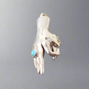 Op de afbeelding: Een zilveren handbedel met een turquoise steen en een gouden accent. De hand houdt een klein, gestileerd dierfiguur vast.
