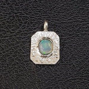 Puede incluir: Un colgante de plata con un borde texturizado y una piedra de ópalo azul claro en el centro.
