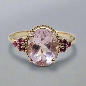 Op de afbeelding: Een gouden ring met een grote ovale roze edelsteen omgeven door een decoratieve rand. De ring is aan beide kanten van de hoofdsteen versierd met kleine roze edelstenen.