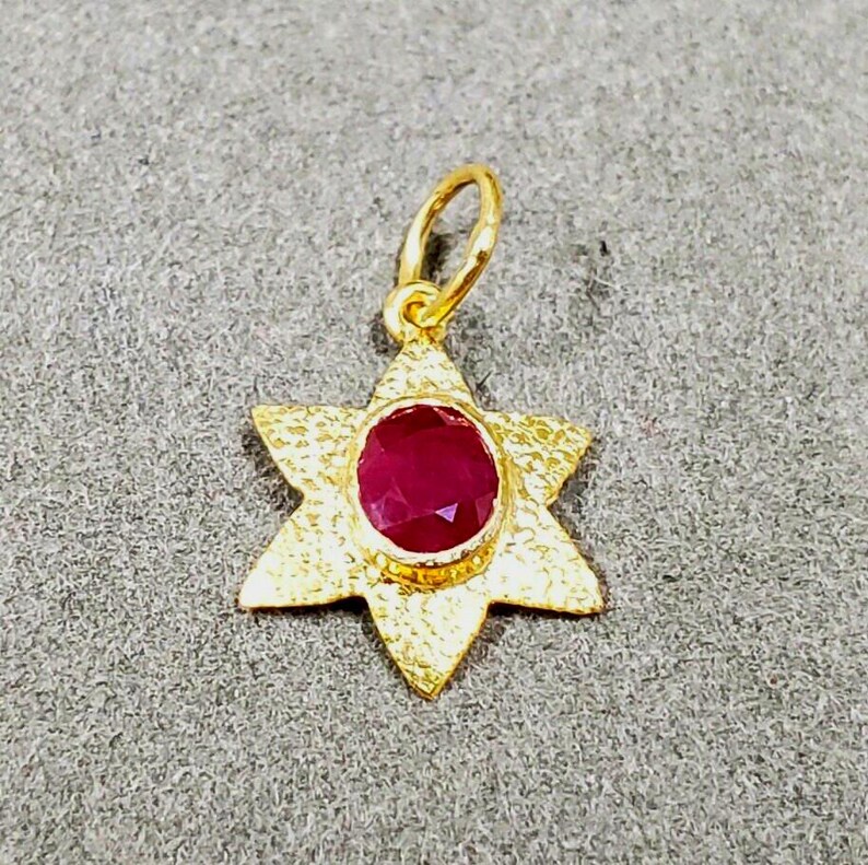 May include: A gold-colored pendant in the shape of a six-pointed star, with a textured surface. The center of the star features a round, deep red gemstone. The pendant has a small loop at the top for a chain.