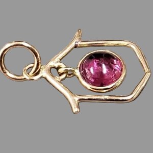 May include: A gold-coloured pendant with a geometric design. A round, pink gemstone is suspended within a hexagonal frame. The pendant has a loop for a chain. The background is a solid grey.