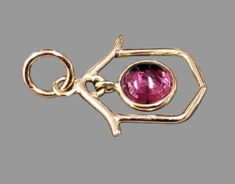 May include: A gold-coloured pendant with a geometric design. A round, pink gemstone is suspended within a hexagonal frame. The pendant has a loop for a chain. The background is a solid grey.