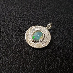 May include: A silver pendant with a round, textured frame and a light blue opal gemstone in the center.