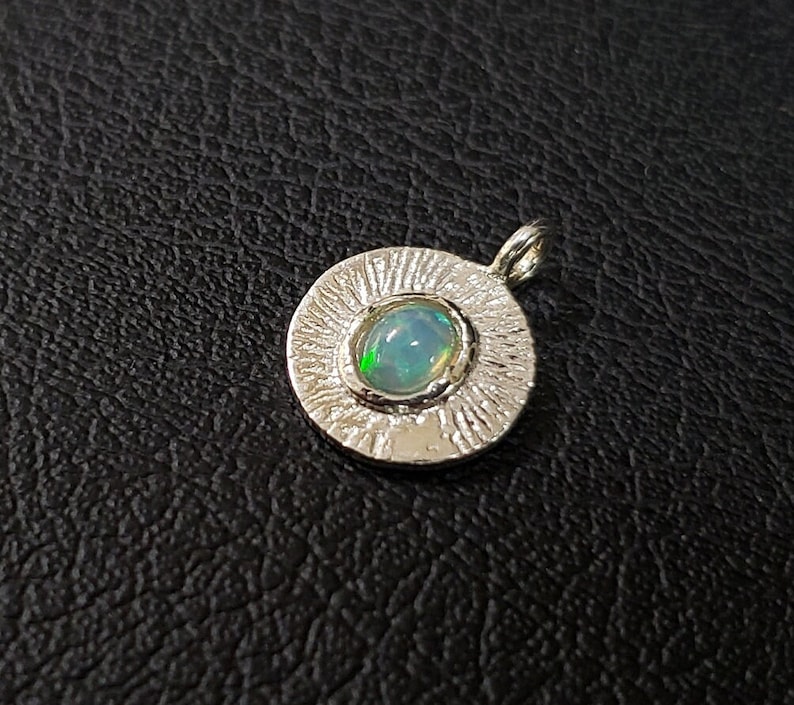 May include: A silver pendant with a round, textured frame and a light blue opal gemstone in the center.