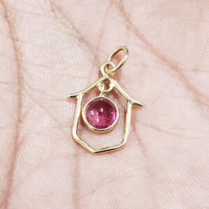 May include: A gold pendant featuring a house-shaped frame. A round, pink gemstone is set in the centre. The pendant has a small loop at the top for a chain. The pendant is likely made of gold.