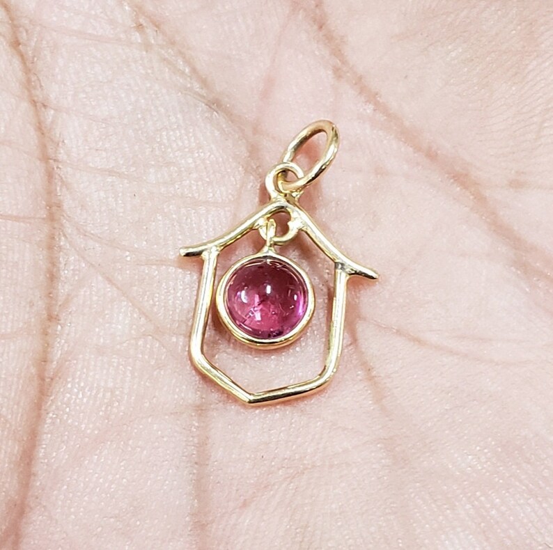 May include: A gold pendant featuring a house-shaped frame. A round, pink gemstone is set in the centre. The pendant has a small loop at the top for a chain. The pendant is likely made of gold.