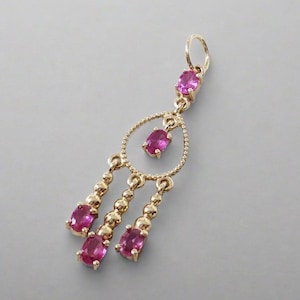 Op de afbeelding: Een goudkleurige hanger met roze edelstenen. De hanger heeft een cirkelvormig ontwerp met een centrale edelsteen en drie bungelende strengen, elk met een edelsteen aan het uiteinde. De bovenkant van de hanger heeft een lus om op te hangen.