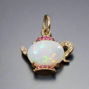 Puede incluir: Un colgante de tetera de oro con una piedra de ópalo blanco y detalles en rosa. El colgante está engastado con pequeñas piedras rosas alrededor del ópalo y el pico de la tetera.