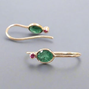 May include: A pair of gold-colored earrings. Each earring features a large, oval-shaped, green gemstone and a smaller, round, pink gemstone. The earrings have a curved design, with a hook for securing them to the ear.