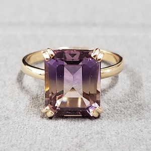 May include: A gold ring featuring a large, rectangular-cut amethyst gemstone. The amethyst displays a gradient of colors, transitioning from a deep purple to a lighter, more translucent hue. The ring's band is a simple, polished gold.