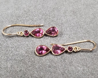 May include: A pair of gold-coloured drop earrings with pink gemstones. Each earring has a small round gem, a larger pear-shaped gem, and a smaller round gem. The earrings are displayed on a grey surface.