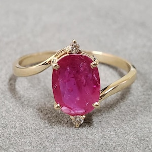 May include: A gold ring featuring a large oval ruby gemstone and smaller diamond accents. The ring has a twisted band.