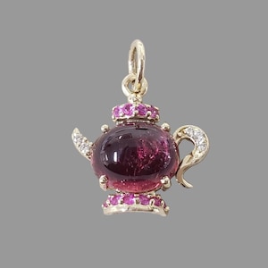 May include: A gold-colored teapot pendant with a deep purple gemstone body. The pendant features pink gemstones on the lid and base, and white gemstones on the handle and spout. The pendant is designed to be worn as jewelry.