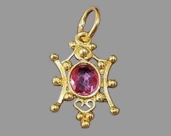 May include: A gold-colored pendant with a central oval-shaped pink gemstone. The pendant has an ornate design with small, round embellishments and a loop at the top for a chain. The pendant is approximately 1 inch in length.