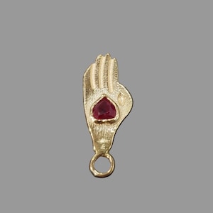 May include: A gold-toned pendant shaped like an open hand with textured fingers and palm details. A single, faceted, heart-shaped red gemstone is set in the centre of the palm. A small, circular jump ring is attached to the base of the hand pendant.