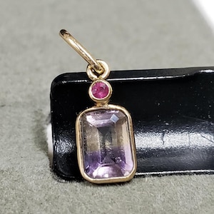 May include: A gold pendant featuring a purple amethyst gemstone and a small red ruby accent.