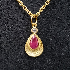Op de afbeelding: Een gouden ketting met een hanger in de vorm van een traan, met een rode edelsteen en een kleinere witte edelsteen.