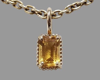 Op de afbeelding: Een goudkleurige ketting met een rechthoekige, amberkleurige hanger. De hanger is gezet in een decoratief frame met een kralenrand en hangt aan een ketting met grote, ovale schakels. De achtergrond is effen grijs.
