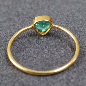 May include: A gold ring with a heart-shaped, light teal gemstone. The ring band is thin and polished, and the gemstone is set in a gold bezel. The ring is displayed on a gray surface.