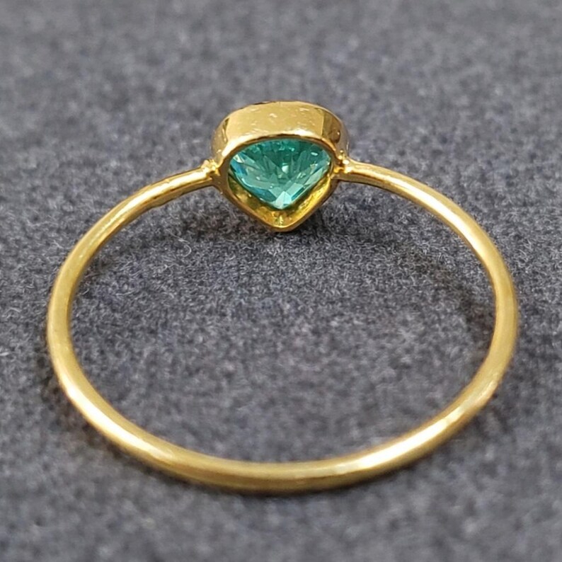 May include: A gold ring with a heart-shaped, light teal gemstone. The ring band is thin and polished, and the gemstone is set in a gold bezel. The ring is displayed on a gray surface.