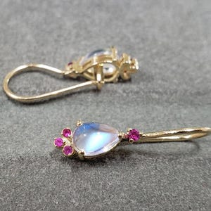 Puede incluir: Un par de pendientes de oro con una piedra de luna en forma de lágrima y piedras preciosas rosas. Los pendientes tienen un diseño simple y elegante.