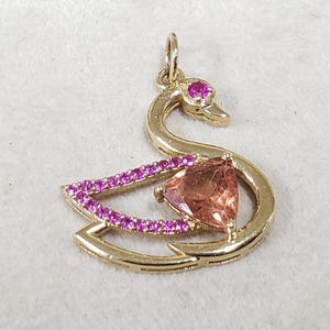 May include: A gold swan pendant featuring a pink gemstone eye and a large, triangular pink gemstone. The swan's wing is adorned with small, pink gemstones. A delicate piece of jewellery.