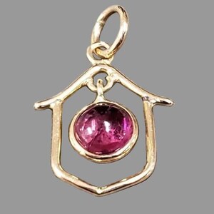 May include: A gold-toned pendant featuring a house-shaped frame, enclosing a round, magenta gemstone. The pendant has a loop for a chain. The gemstone is a striking, vivid magenta colour, adding a pop of colour to the design.