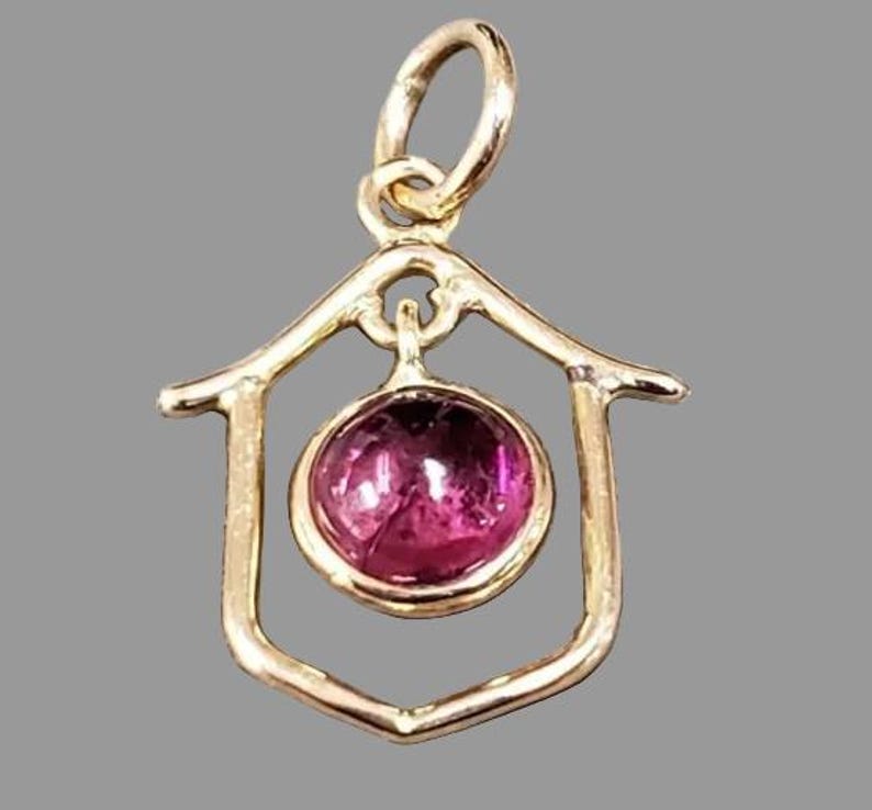 May include: A gold-toned pendant featuring a house-shaped frame, enclosing a round, magenta gemstone. The pendant has a loop for a chain. The gemstone is a striking, vivid magenta colour, adding a pop of colour to the design.
