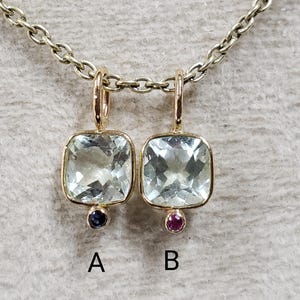 May include: Two gold pendants with square-shaped clear gemstones. The pendant on the left has a dark blue gemstone at the bottom, while the pendant on the right has a pink gemstone at the bottom.