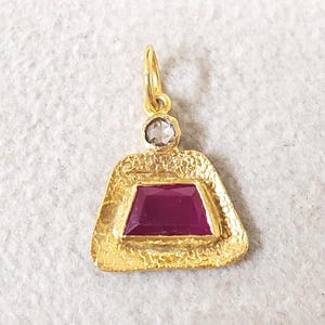 Puede incluir: Colgante de oro con una gema rectangular de color rojo intenso en el centro. El colgante tiene una superficie texturizada y una pequeña gema transparente encima de la piedra roja. Un lazo dorado en la parte superior para colgar.