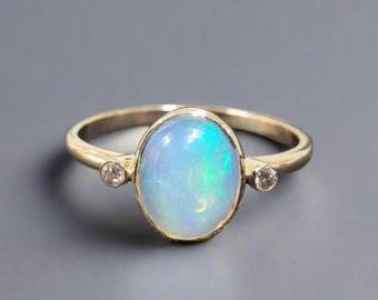 May include: A gold ring featuring a large oval opal gemstone with iridescent blue and green hues. Two small, round accent stones flank the opal. The ring is set against a gray background.