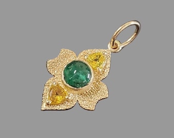 Könnte beinhalten: Goldener Anhänger mit floralem Design. Der Anhänger hat einen großen, runden, grünen Edelstein in der Mitte, mit zwei gelben, dreieckigen Edelsteinen und einer strukturierten Goldoberfläche. Der Anhänger hat eine Schlaufe für eine Kette.