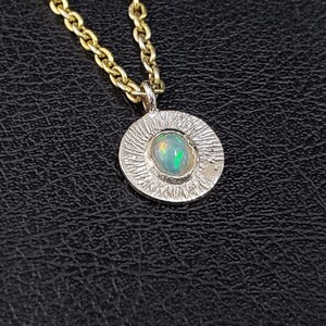 May include: A silver pendant necklace with a textured circular design featuring a green opal gemstone. The necklace is on a gold chain.