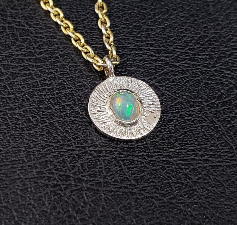 May include: A silver pendant necklace with a textured circular design featuring a green opal gemstone. The necklace is on a gold chain.