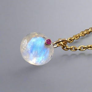 May include: A round, iridescent moonstone pendant with a small, round ruby accent. The pendant is attached to a gold chain necklace. The moonstone has a blue and white glow. Jewellery.