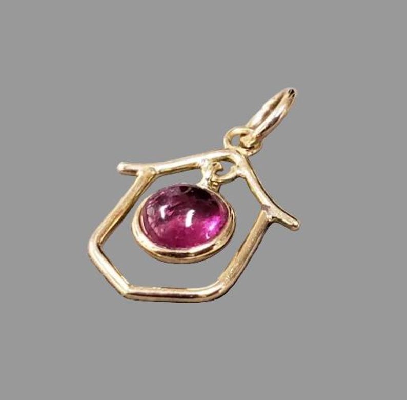 May include: A gold-coloured pendant with a deep pink, round gemstone set in a geometric frame. The pendant has a small loop at the top for a chain. The gemstone is the focal point, with a polished, reflective surface.